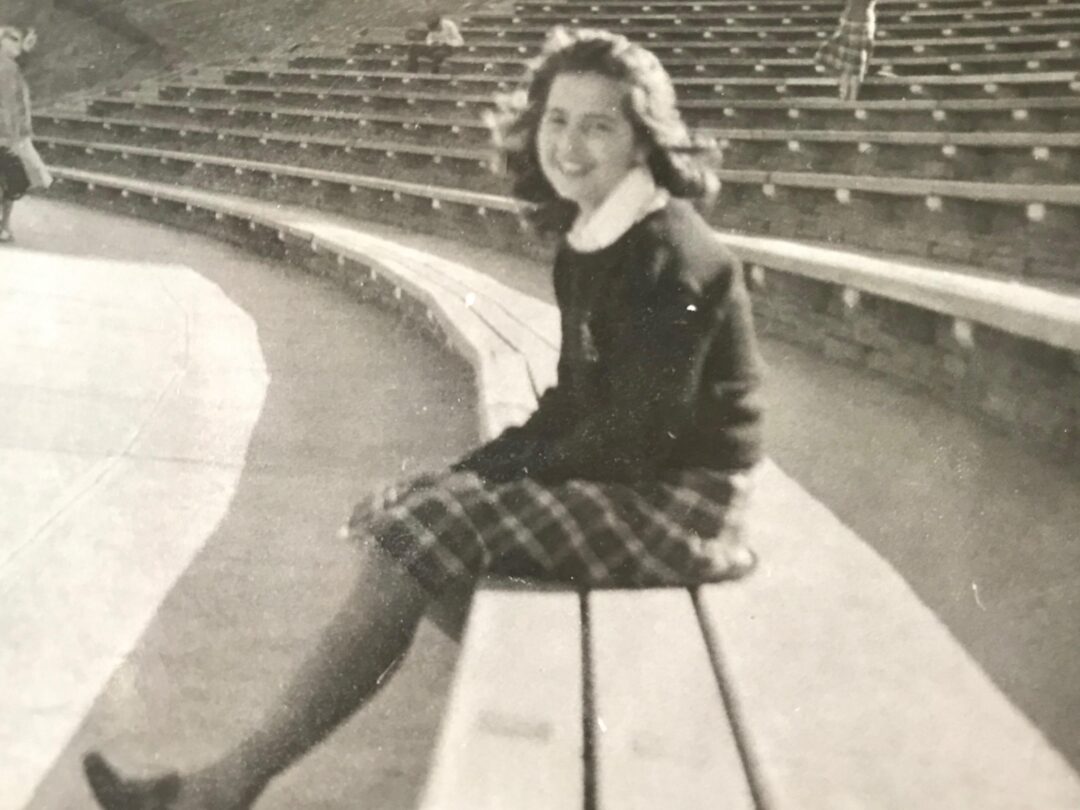 A young woman with wavy hair sits on a wooden bench in an empty outdoor amphitheater, smiling at the camera. She wears a plaid skirt and a dark sweater. The photo is black and white and slightly blurred.