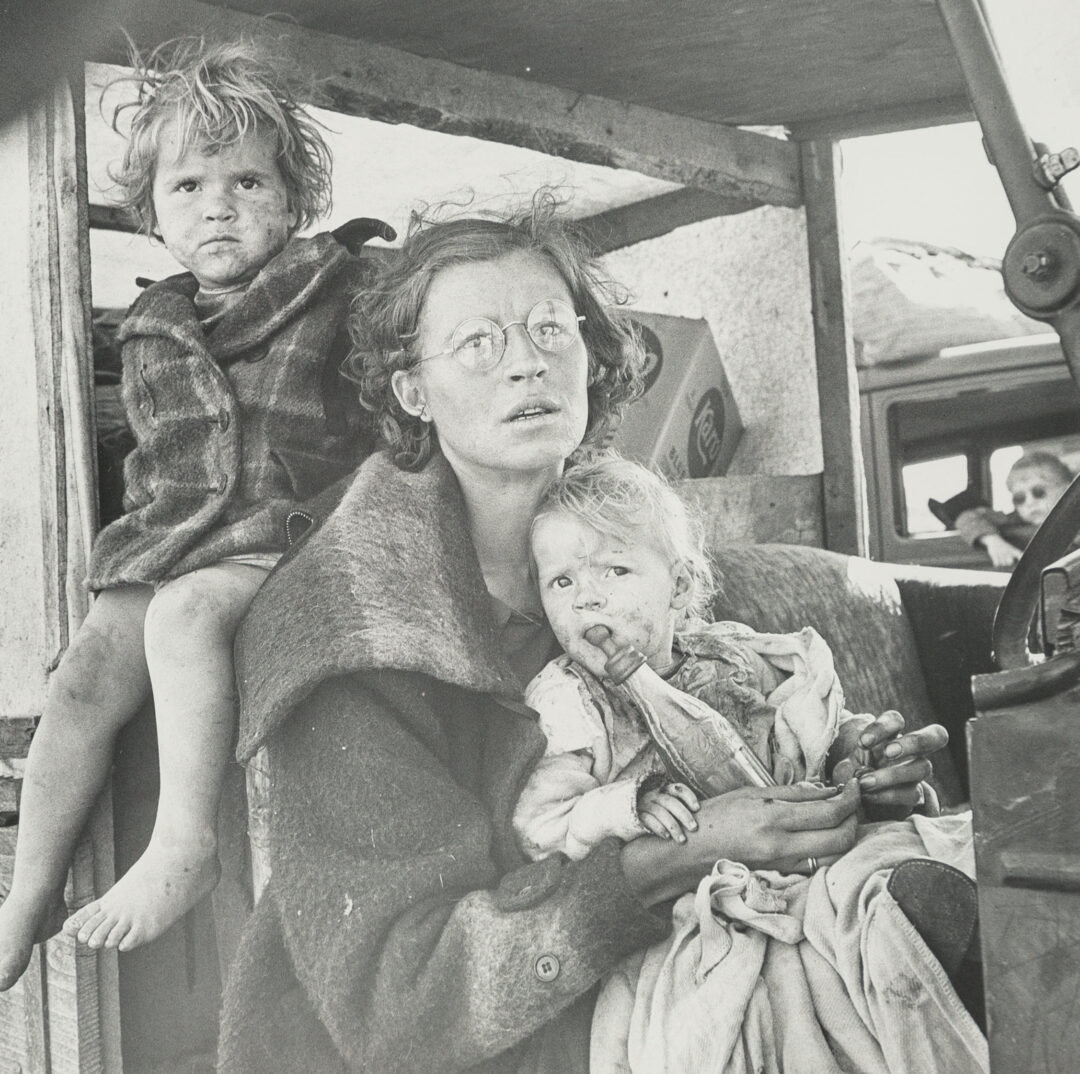 A woman with glasses sits holding a young child with a bottle while another barefoot child sits beside her. All three appear somber and wear worn clothes, framed by a vehicle in a dusty, outdoor setting.