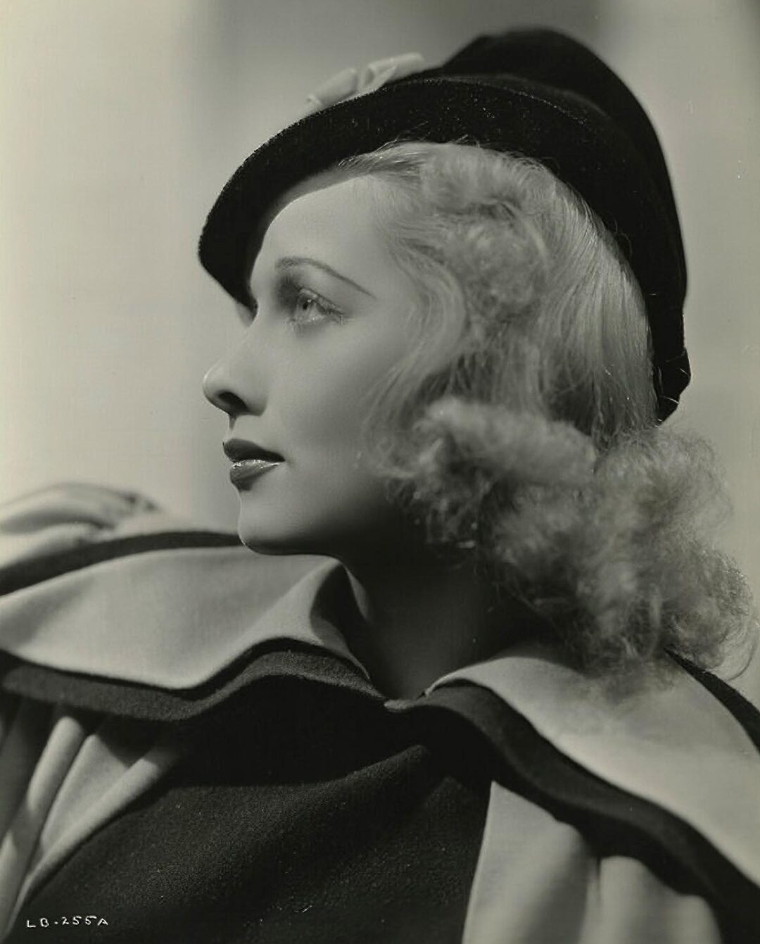 Black-and-white portrait of a woman in profile, wearing a dark hat and a cape with a wide collar. She has wavy, shoulder-length hair, and gazes upward with a serene expression. The lighting creates a soft, elegant mood.