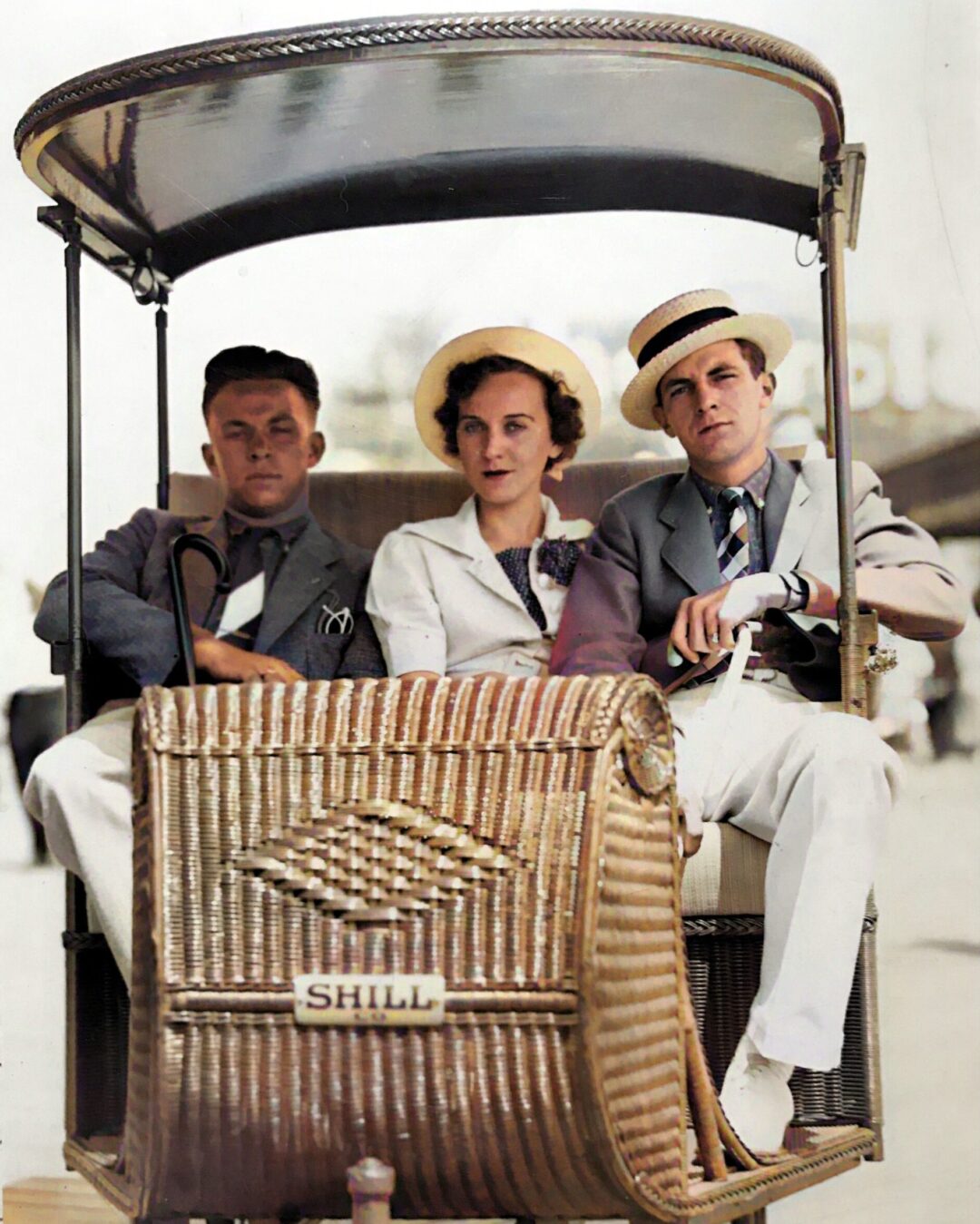 Three people, two men and one woman, sit in a vintage wicker rickshaw labeled “SHILL.” All are dressed in light-colored, elegant 1930s-style clothing and hats, looking directly at the camera.