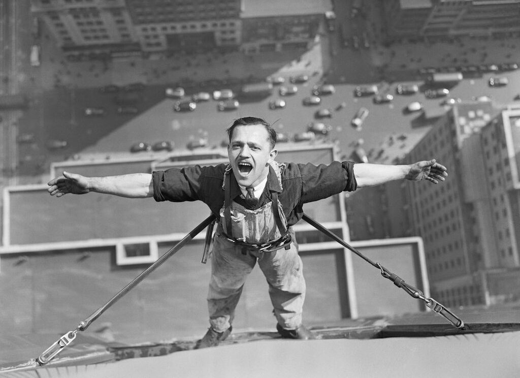 A man in work clothes stands on the edge of a tall building, arms outstretched, secured by safety harnesses, high above a busy city street with cars and pedestrians far below.
