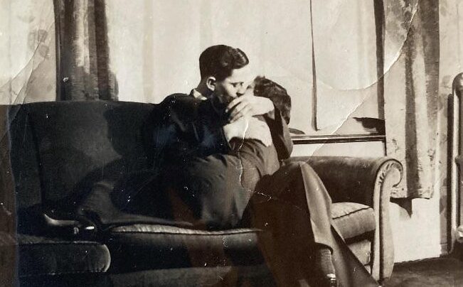 A black-and-white vintage photo shows two people embracing closely on a couch in a sunlit room, with patterned curtains and a radiator visible in the background.