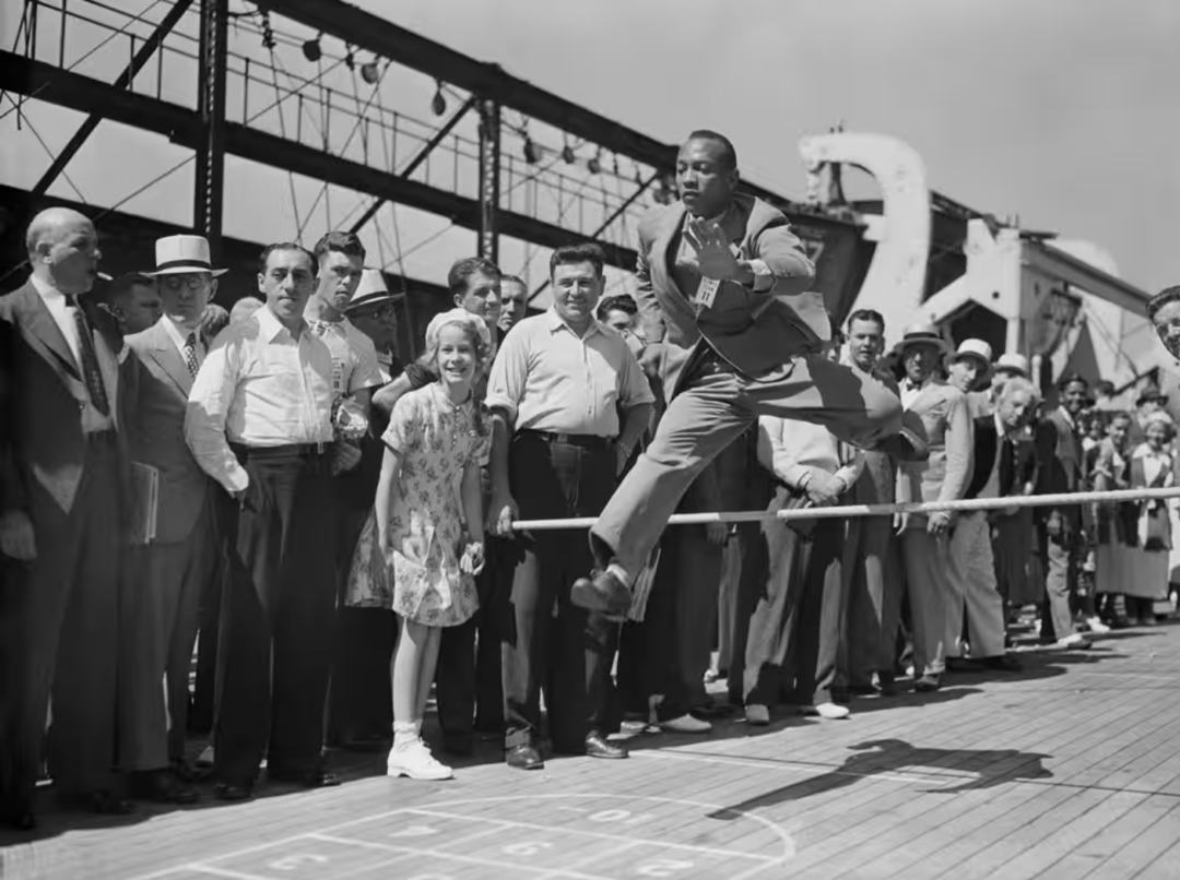 A man in a suit jumps energetically over a shuffleboard court on a ship deck, while a crowd of men and women watches and smiles in the background, under a sunny sky.