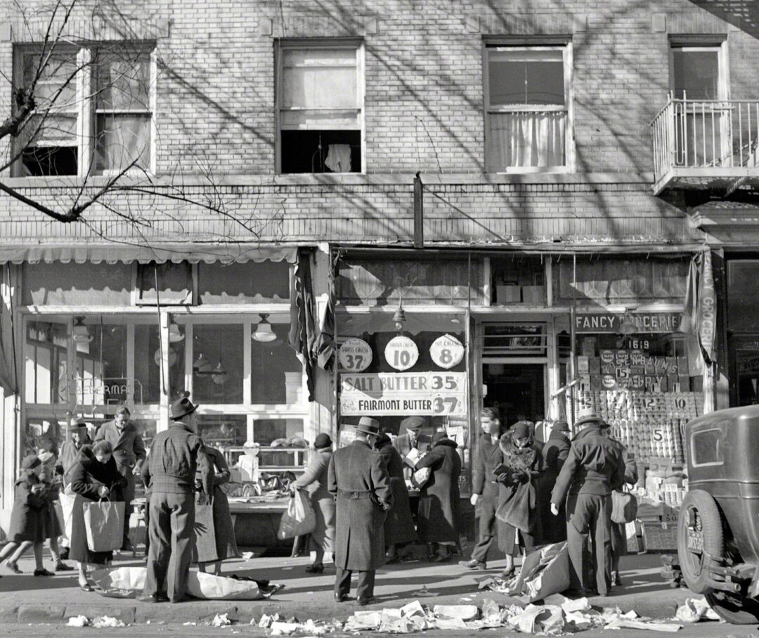 Black-and-white photo of people gathered on a sidewalk outside a grocery store. Some are shopping at outdoor produce stands; others are talking. Signs in the window advertise prices for salt butter and eggs. A car is parked nearby.