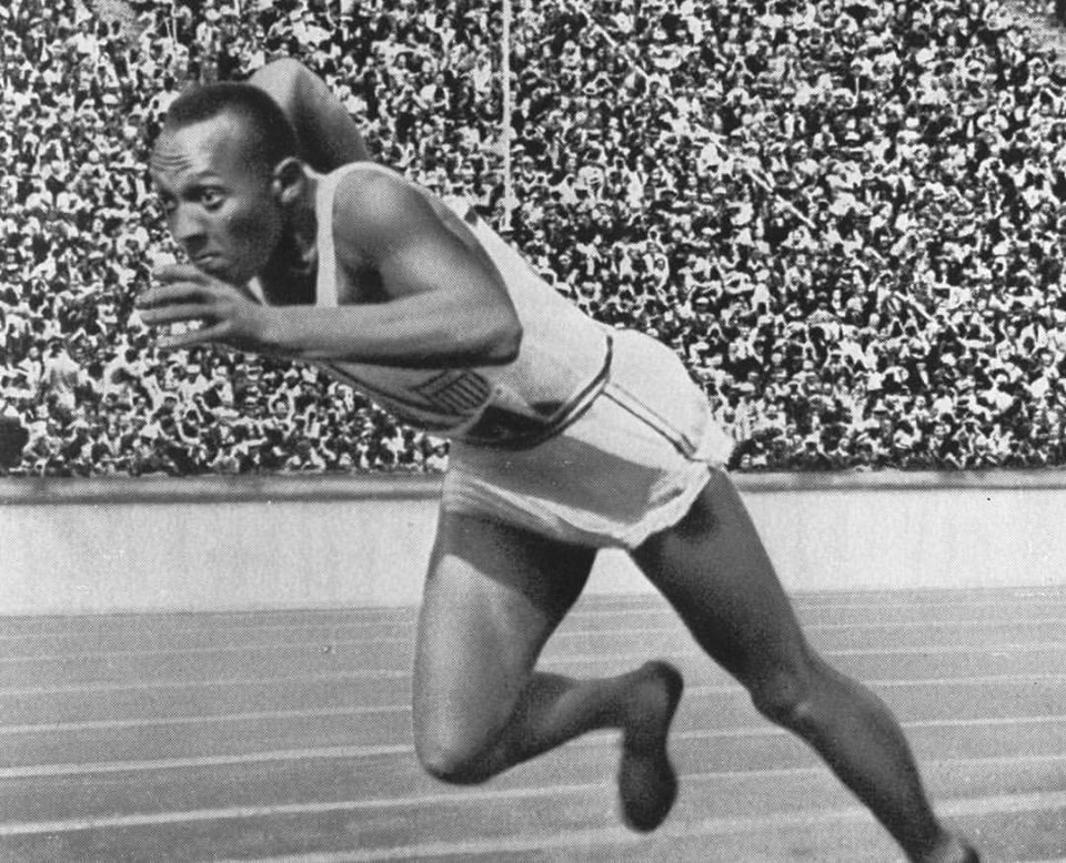 A Black male track athlete in mid-sprint on a running track, with a large crowd in the stadium stands behind him. He wears a sleeveless top and shorts, and appears focused on his race.