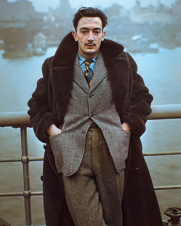 A man with dark hair and a thin mustache stands by a railing, wearing a long dark coat over a checked suit and striped tie. The background is foggy, with water and blurred buildings visible.