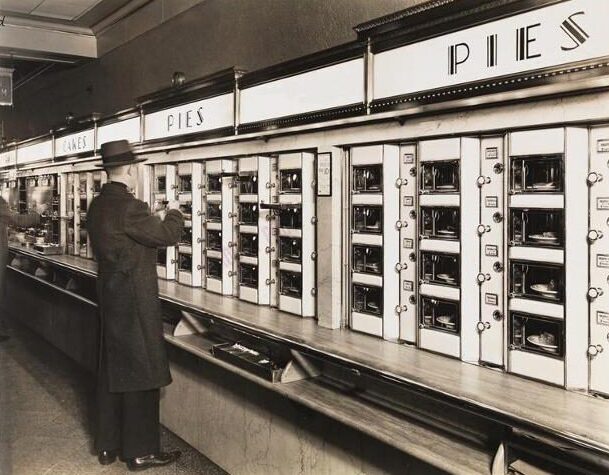 A man in a coat and hat selects food from a vintage automat, with labeled compartments for pies and cakes, each containing plates of pastries behind glass doors.