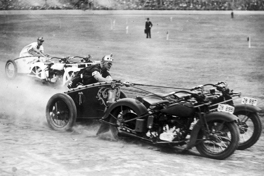 Three people in vintage motorcycles with sidecars race around a dusty dirt track, stirring up clouds behind them, while a small crowd watches in the background.