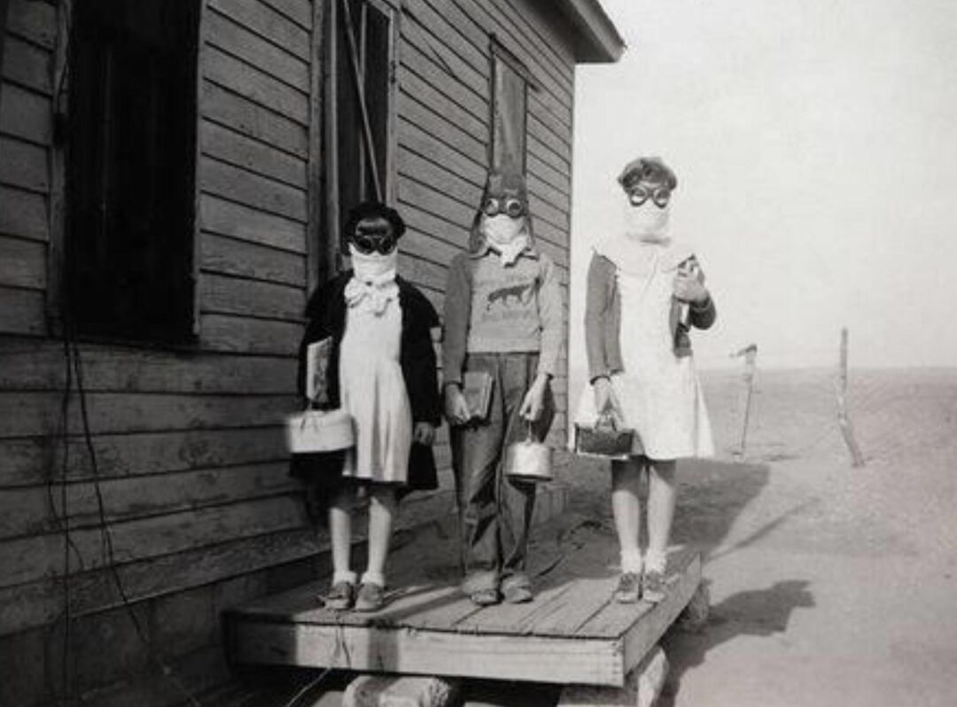 Three children wearing masks and goggles stand on a wooden platform outside a building, each holding books and lunch containers. The background shows a barren, dusty landscape.