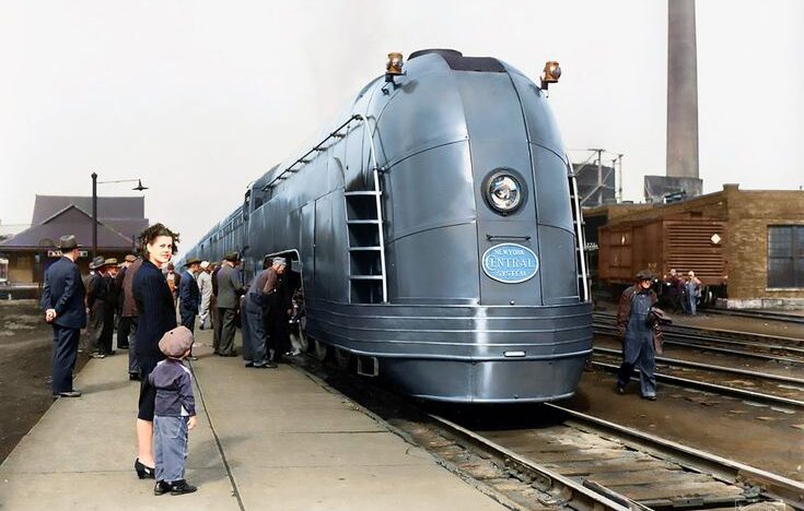 A sleek, streamlined 1930s passenger train at a station platform, with people boarding and onlookers, including a woman and child, standing nearby. The scene appears to be from a past era.