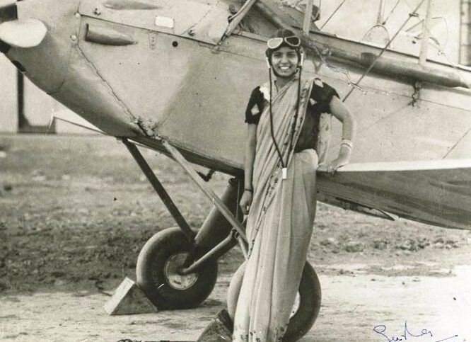 A woman in a sari, wearing aviator goggles and smiling, stands beside a vintage propeller airplane. The black-and-white photo appears historical, and the woman is posing confidently near the wing and landing gear.