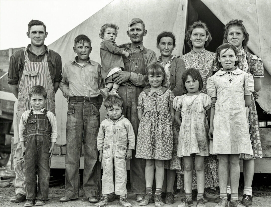 A black-and-white photo of a large family—two adults and ten children—posing in front of a canvas tent during the Great Depression era. The children are wearing worn clothes and most have serious expressions.