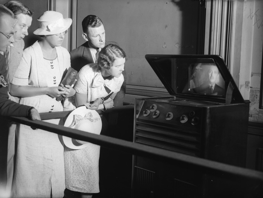 A group of people in 1930s attire closely watch an early television set displaying a man's face. The onlookers appear intrigued and focused, with one woman holding a hat and leaning in for a better view.