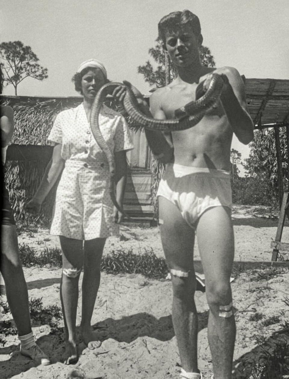 A shirtless man in swim trunks holds a large snake, standing next to a woman in a polka-dot romper and hat outdoors, with another person partially visible on the left.