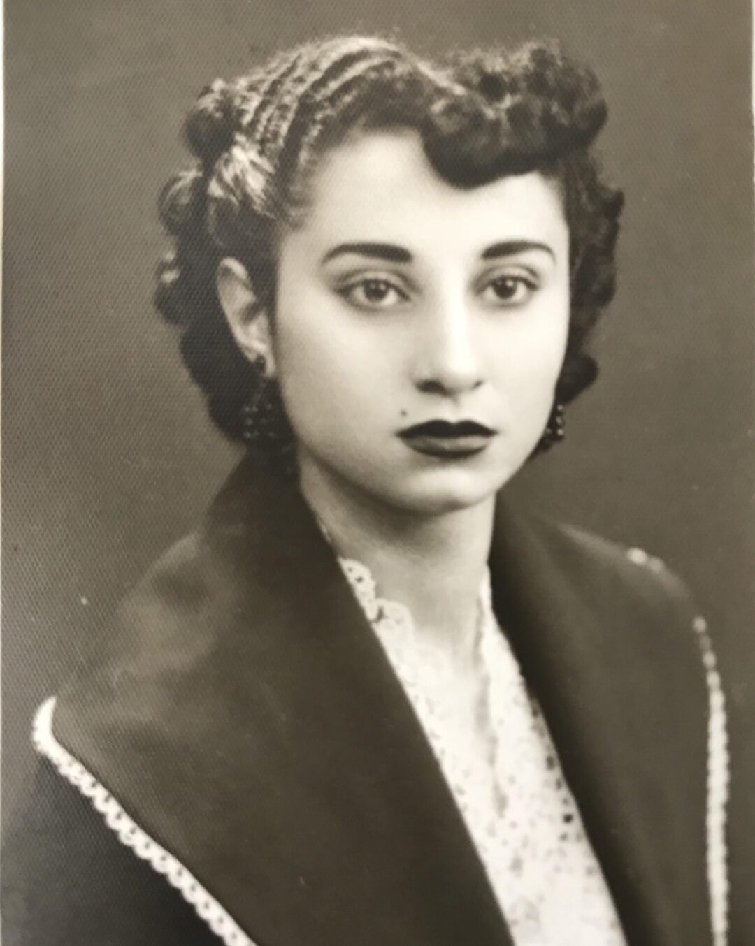 Black and white portrait of a young woman with short, curled hair, dark lipstick, and earrings. She is wearing a shawl with a decorative edge over a lace blouse and is looking directly at the camera with a neutral expression.