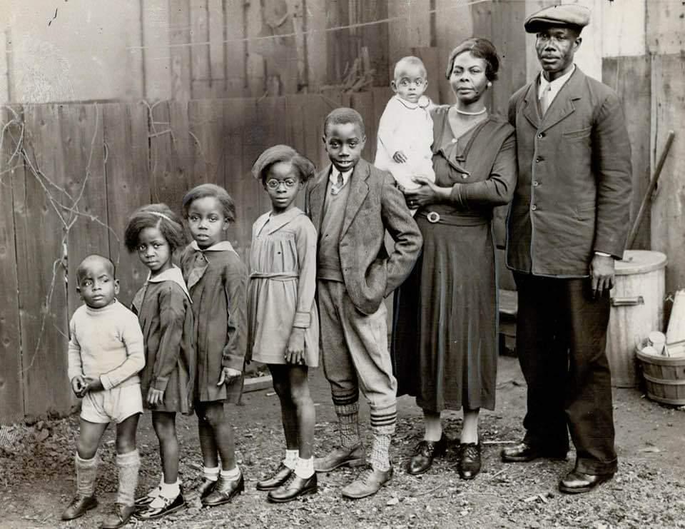 A Black family of seven, including five children and two adults, stands outdoors in front of a wooden fence. The adults are dressed formally, and the children wear a variety of dresses and shorts. One adult holds a baby.