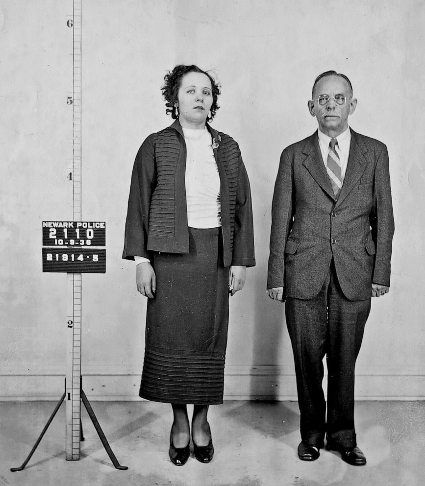 A woman and a man stand side by side for a police photo in formal clothing. A sign beside them reads "Newark Police" with numbers and a date, suggesting a mugshot or police identification photo.