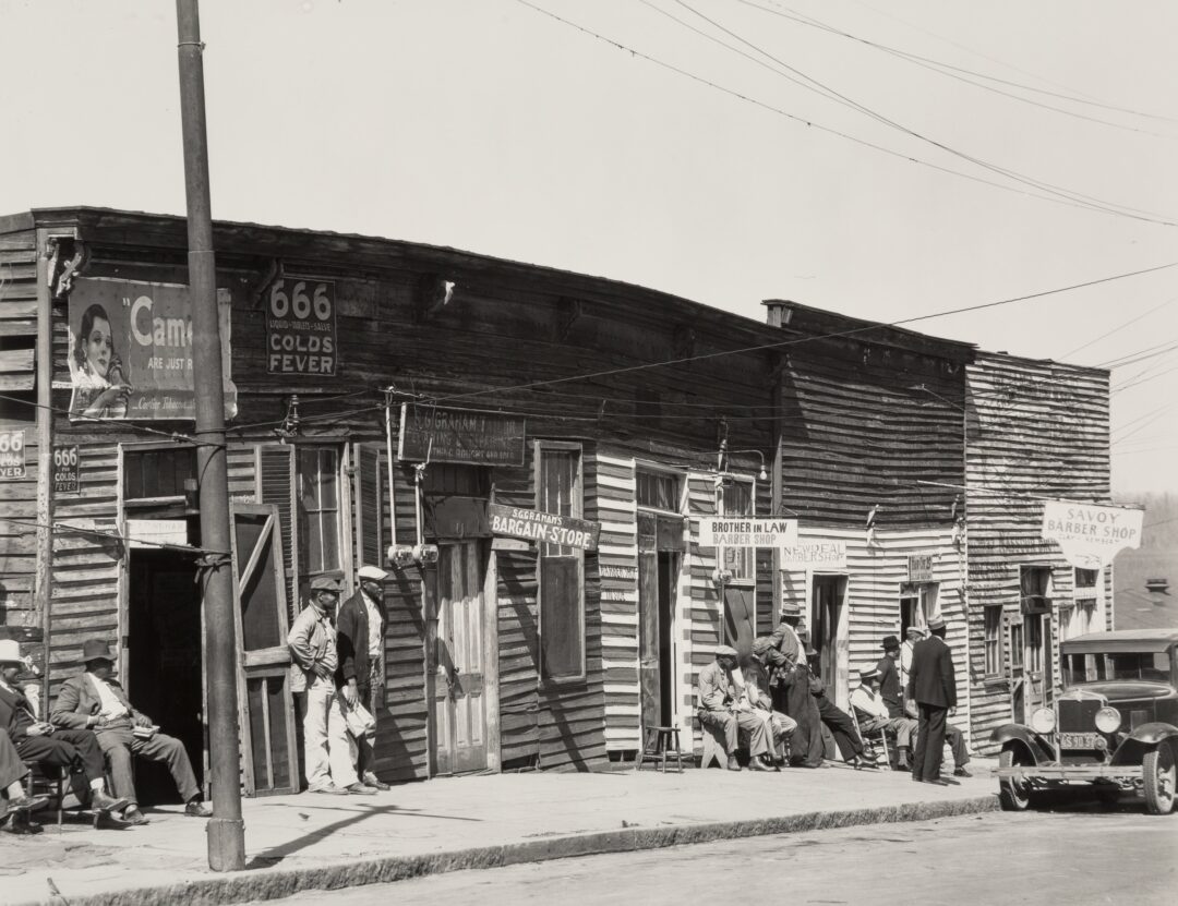 Black and white photo of a row of worn wooden shops on a street, with several people sitting and standing outside. Signs advertise a bargain store, barber shop, and cold remedy. An old car is parked in front.