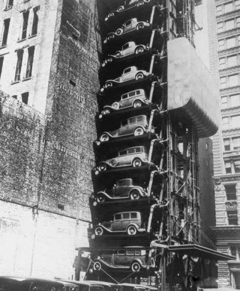 Black and white photo of a vertical car parking tower, with multiple vintage cars stacked on mechanical platforms between tall buildings in a city.