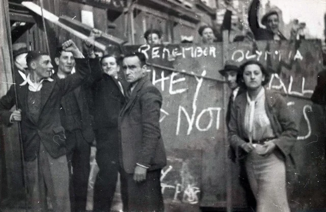 A group of people, some raising fists, stand in front of a barricade with handwritten slogans. The scene appears tense, with men and women gathered closely, suggesting a protest or demonstration.