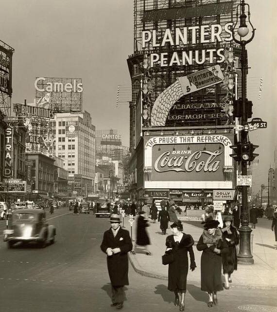 Black-and-white photo of a busy city street in the 1940s with large vintage advertisements for Planters Peanuts, Coca-Cola, and Camels. People walk on the sidewalk and old-fashioned cars drive by.