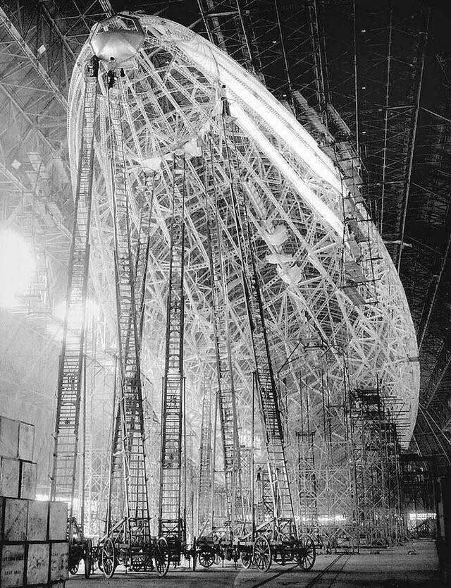 A large airship is under construction inside a vast hangar. Numerous scaffolds, ladders, and workers surround the airship’s skeletal frame, highlighting its immense size and intricate structure.