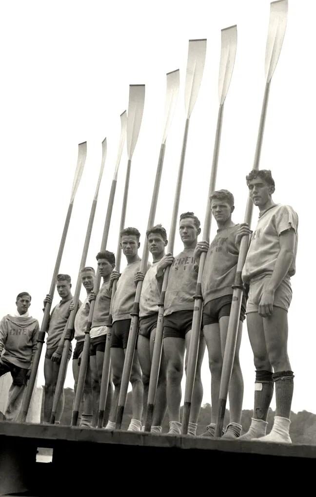 Nine young men stand in a row on a dock, each holding a tall rowing oar upright. They wear athletic shirts and shorts, and are outdoors with trees visible in the background.
