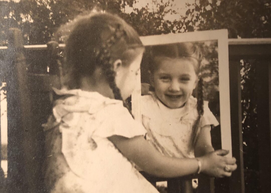 A young girl with braided hair looks into a mirror outside, smiling at her reflection. The photo is sepia-toned, giving it a vintage feel. She wears a light-colored dress and stands near a wooden railing.