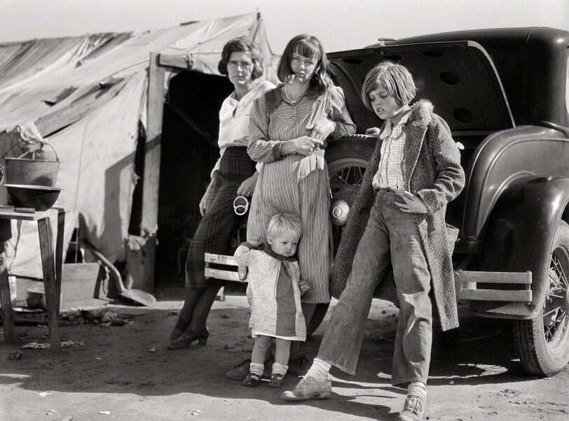 Four children stand in front of an open car trunk and a canvas tent, wearing worn clothes. One small child stands close to an older girl as the others look toward the camera, evoking a sense of hardship during the Great Depression.