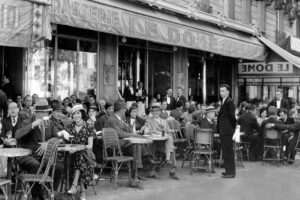 Black and white photo of people in 1920s-style clothing sitting at small tables outside a busy Parisian café, with waiters serving customers and a sign reading “Le Dome” above the entrance.