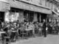 Black and white photo of people in 1920s-style clothing sitting at small tables outside a busy Parisian caf&eacute;, with waiters serving customers and a sign reading &ldquo;Le Dome&rdquo; above the entrance.