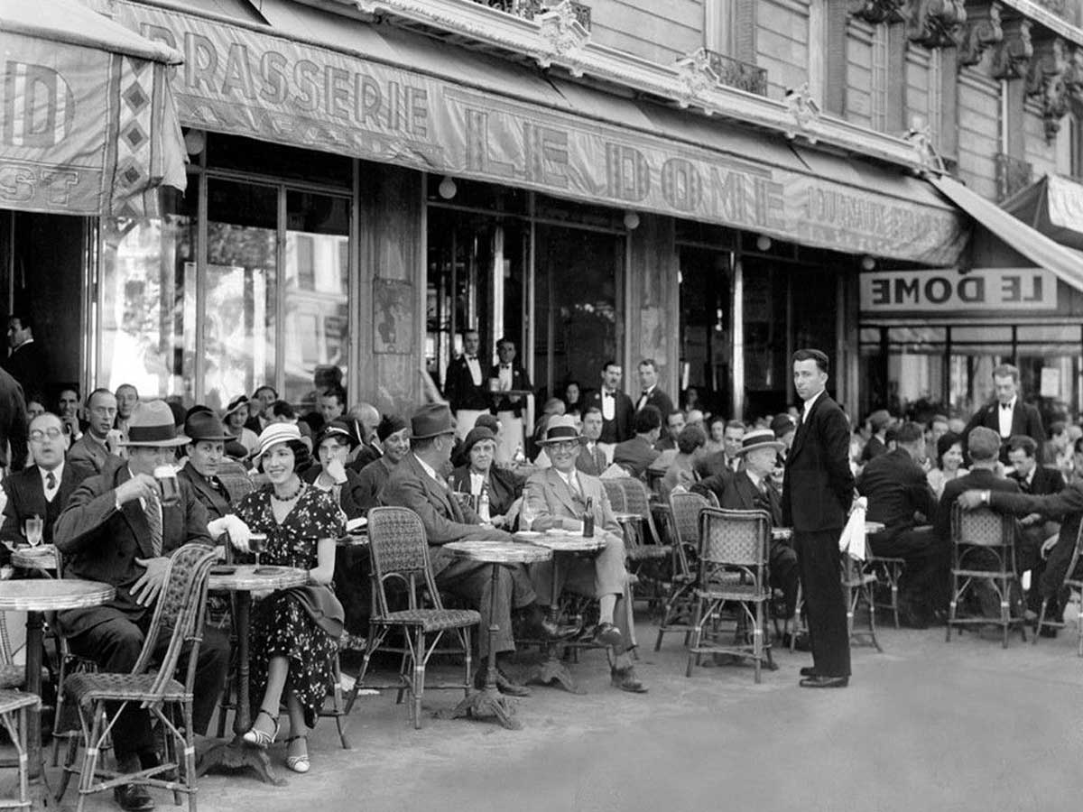 Black and white photo of people in 1920s-style clothing sitting at small tables outside a busy Parisian café, with waiters serving customers and a sign reading “Le Dome” above the entrance.