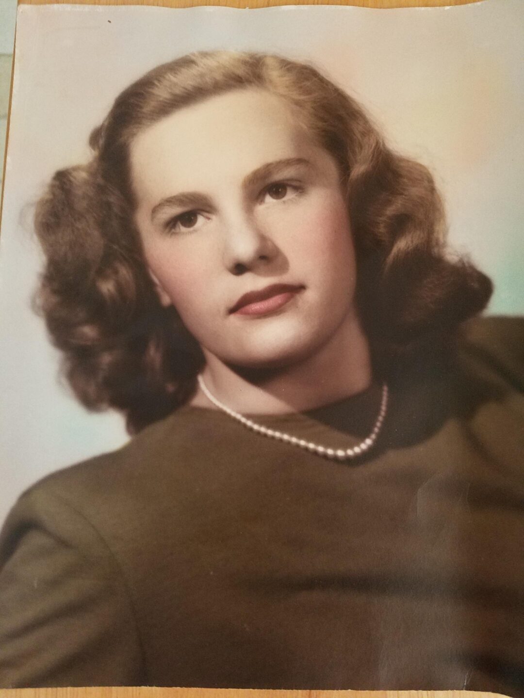 A vintage portrait of a young woman with wavy brown hair, wearing a dark top and a single-strand pearl necklace, looking slightly off camera with a neutral expression against a softly colored background.