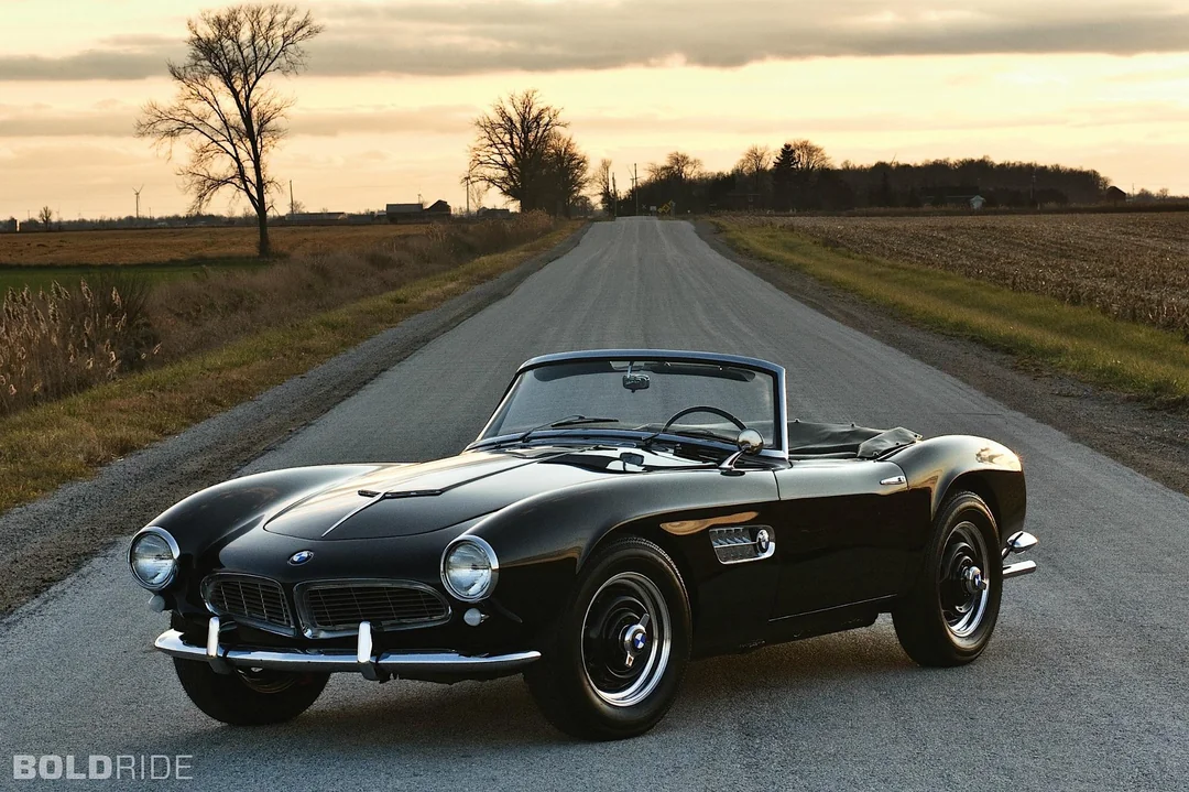 A classic black convertible BMW sports car is parked on a rural road at sunset, surrounded by open fields and distant trees under a cloudy sky.