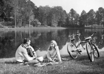 A young man and woman sit on a picnic blanket by a lake, smiling at each other. Two bicycles are parked nearby on the grass, with trees and calm water in the background.