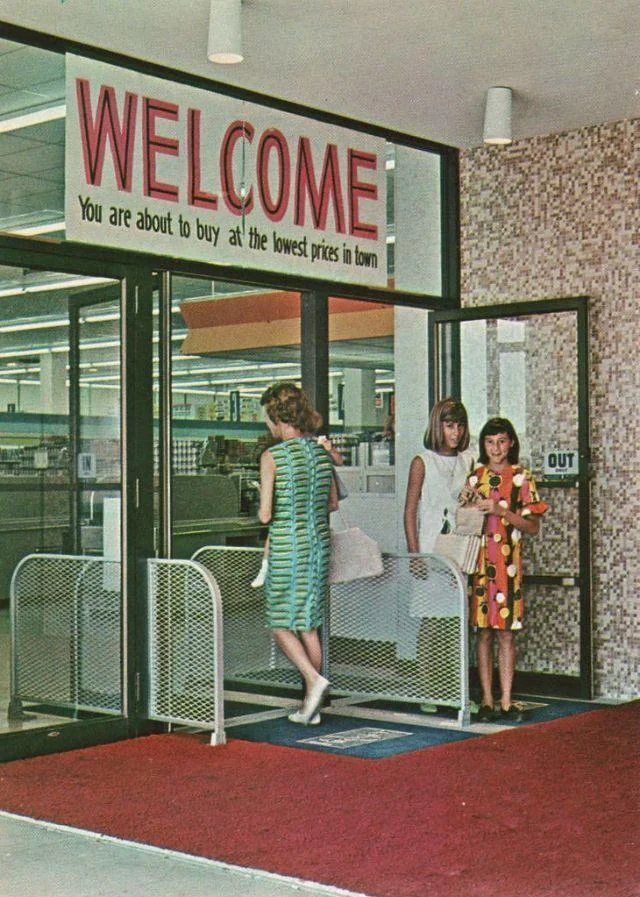 Three women in colorful 1960s dresses enter a store through glass doors under a large sign reading "WELCOME. You are about to buy at the lowest prices in town." The entrance has a red carpet and a patterned wall.