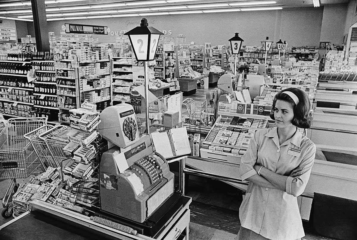 A black-and-white photo of a woman standing with folded arms in front of old-fashioned cash registers in a busy supermarket, surrounded by grocery items, shopping carts, and shelves stocked with products.