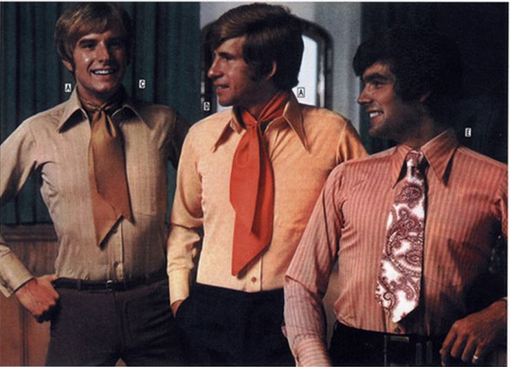 Three men stand together, smiling and wearing 1970s-style shirts with wide collars and bold, colorful neckties. The neckties vary in style, including large, solid-colored, and patterned designs.