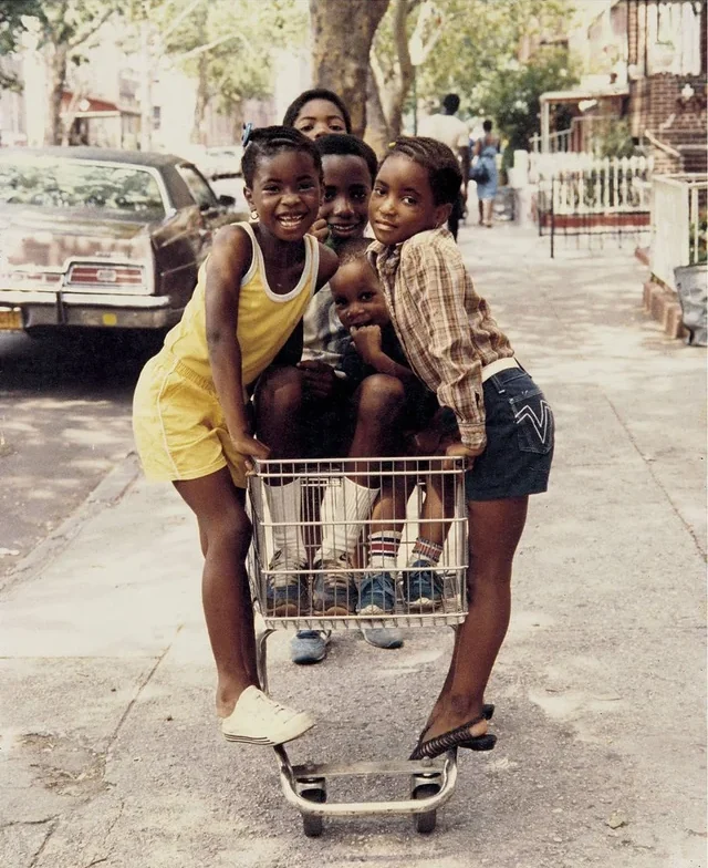 Five smiling children stand and sit together on a shopping cart on a sunny city sidewalk, with trees, parked cars, and apartment buildings in the background.