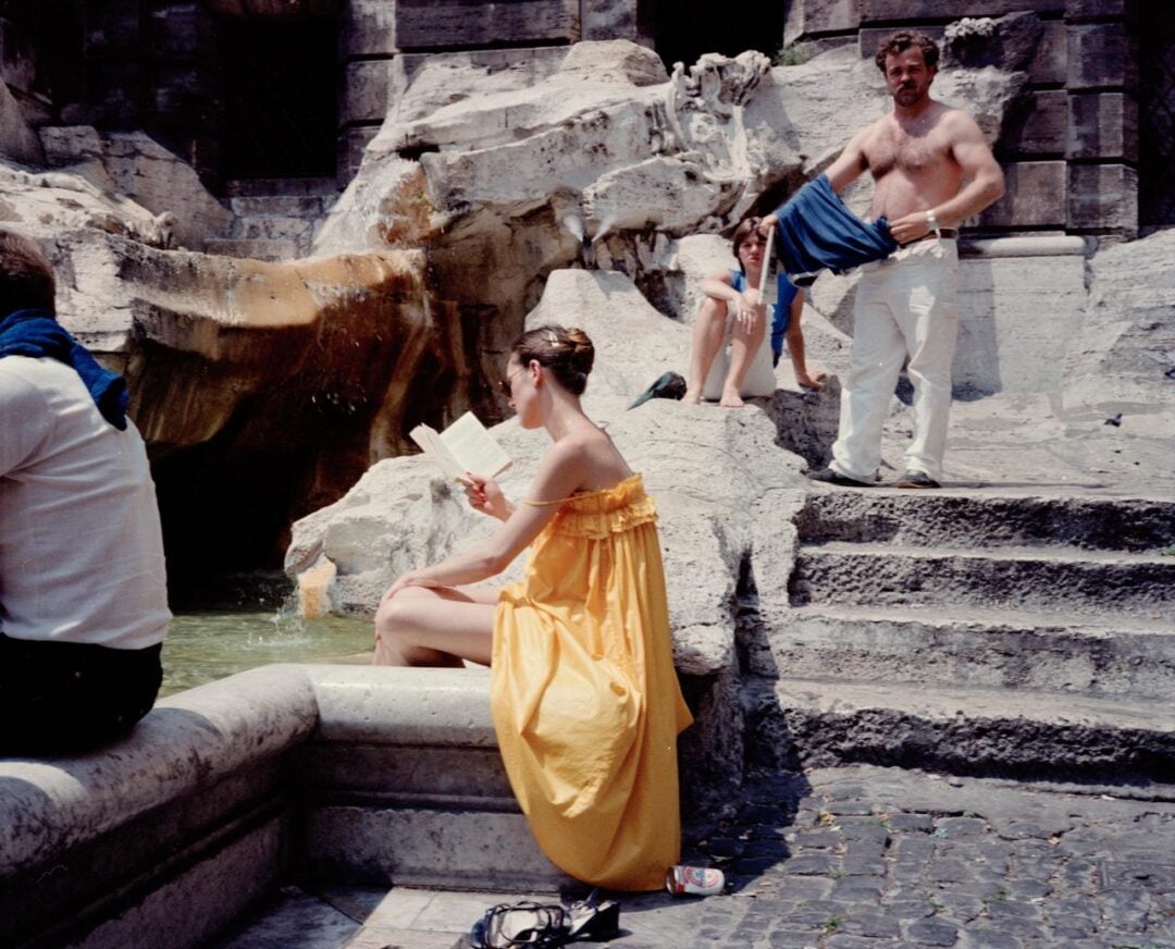 A woman in a bright yellow dress sits reading beside a fountain, while two people relax on nearby steps—one shirtless and another in swimwear. Empty sandals and a drink can rest on the ground. The scene is sunlit and casual.