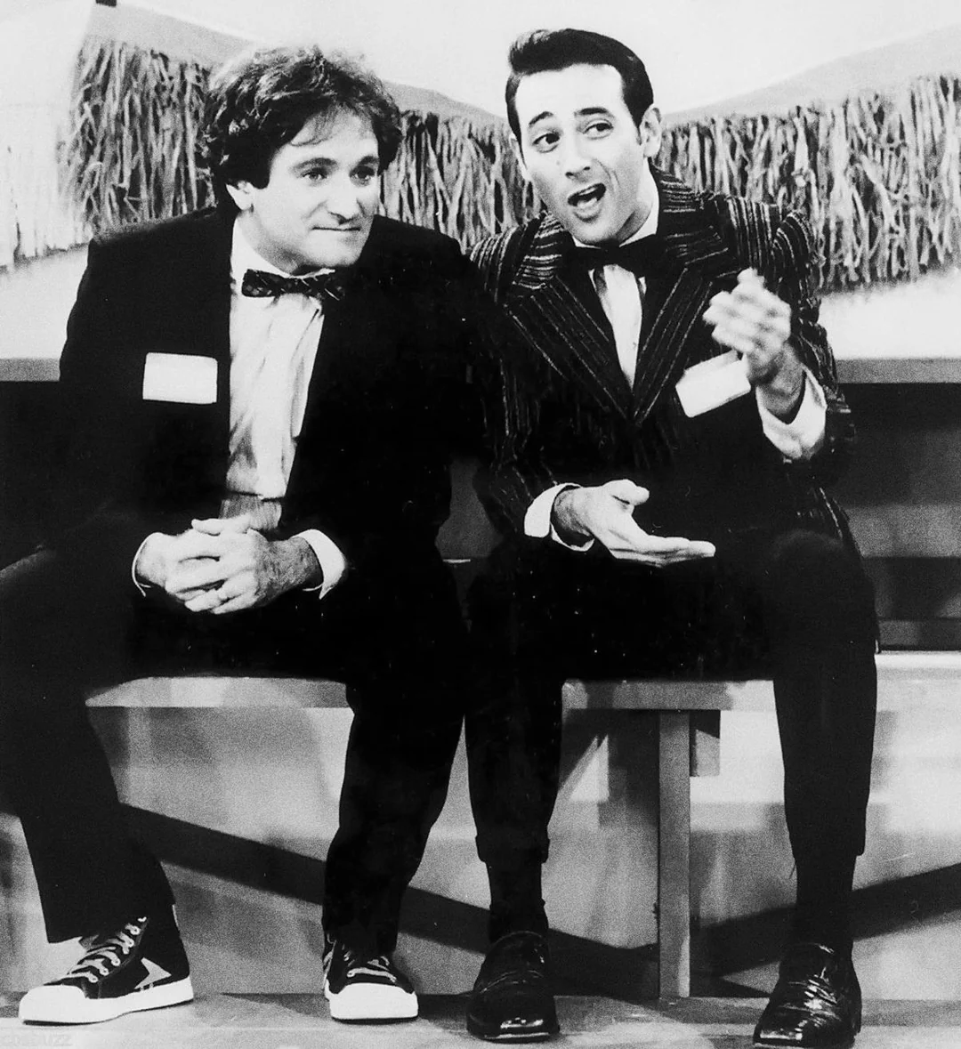 Two men in suits sit on a bench. The man on the left wears a bow tie and mismatched sneakers, while the man on the right gestures animatedly. Both appear to be mid-conversation on a stage with decorative fringe behind them.