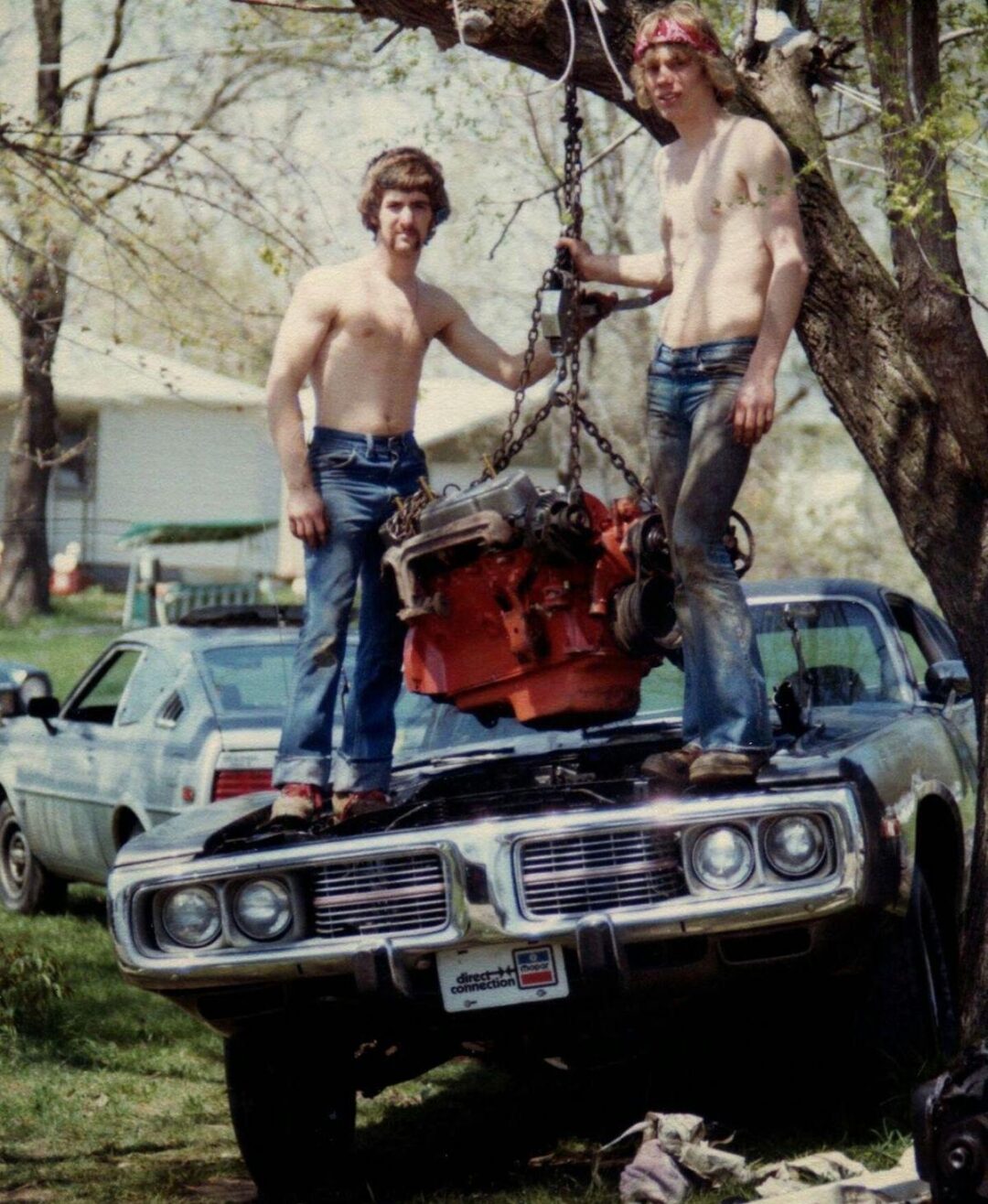Two shirtless men in jeans stand on the hood of an old car, using a chain hoist to lift an engine out of the vehicle. There are trees, a house, and other cars visible in the background.