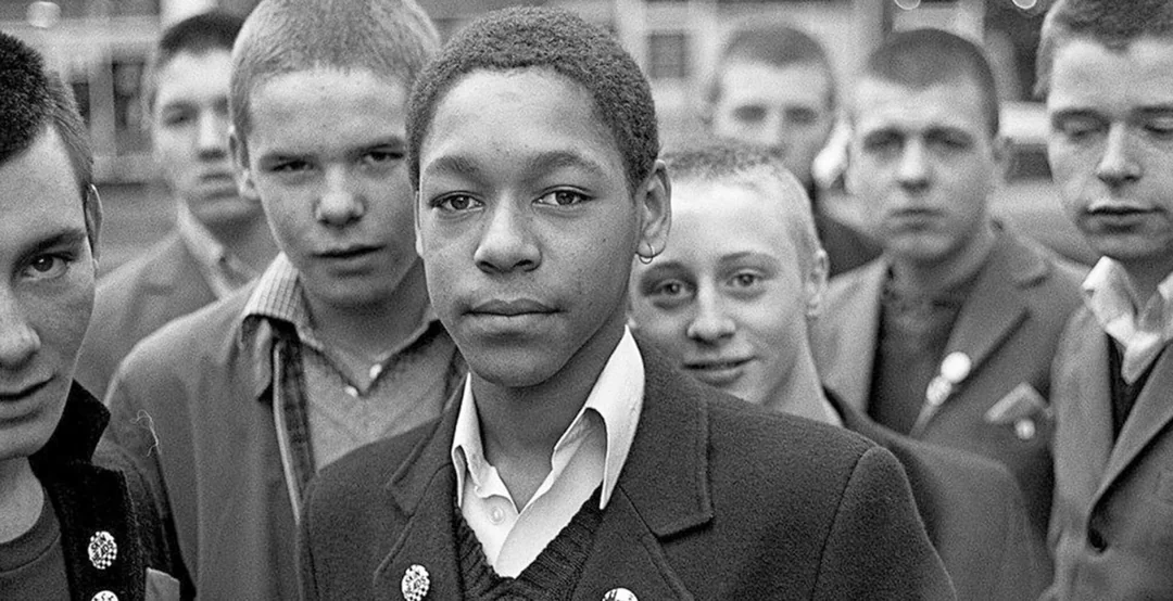 A group of young boys in jackets stand close together, looking at the camera. The central boy wears a white shirt and has a calm expression. The photo is black and white, with a background of blurred buildings.