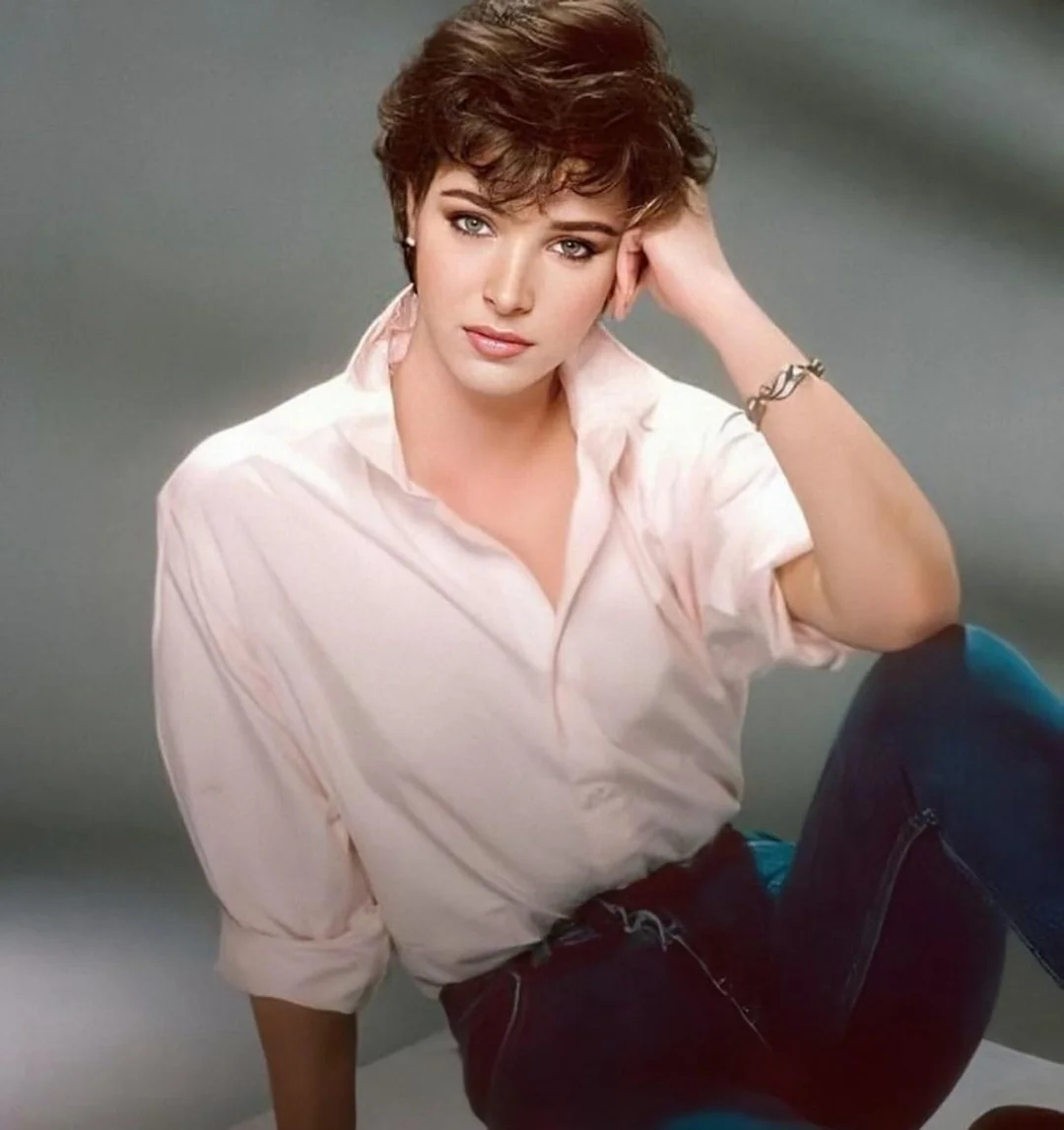 A person with short, wavy brown hair and light makeup sits casually, wearing a loose pale pink shirt and blue jeans, with one arm resting on their knee and the other hand touching their head. The background is softly blurred.