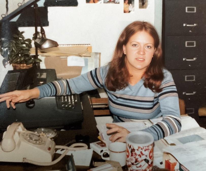 A woman with long brown hair sits at a cluttered desk with papers, a rotary phone, a typewriter, a mug, and office supplies. She wears a striped sweater and looks toward the camera, in a vintage office setting.