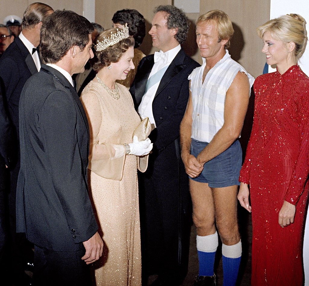 A woman in a tiara and elegant dress greets people at a formal event. A man in a sleeveless shirt, shorts, and boots stands next to a woman in a sparkling red gown. Others in suits are visible in the background.