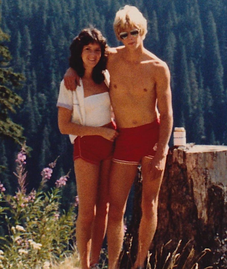 A woman and a shirtless man in red shorts pose together outdoors in front of a tree stump, with a forested mountain slope in the background. The man has his arm around the woman's shoulders.