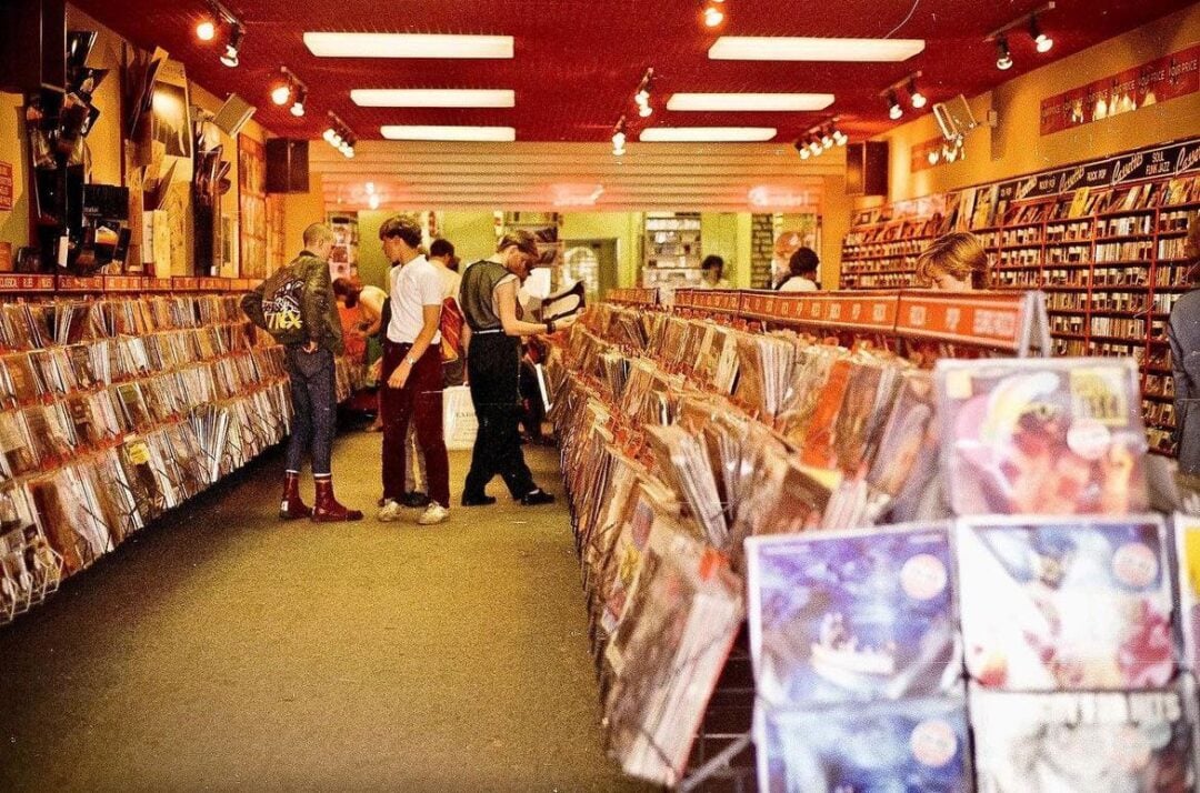 People browse vinyl records in a retro-style record store with rows of album covers on display, bright lighting, and shelves lining the walls filled with more records and music items.