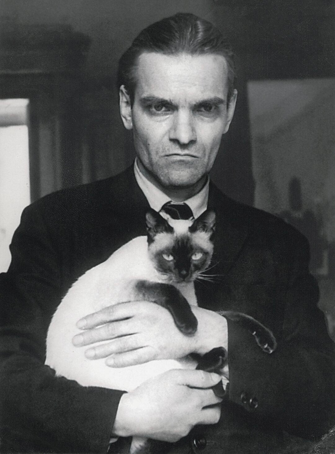 A serious-looking man in a dark suit holds a Siamese cat in his arms. The photo is black and white, and the background is blurred.