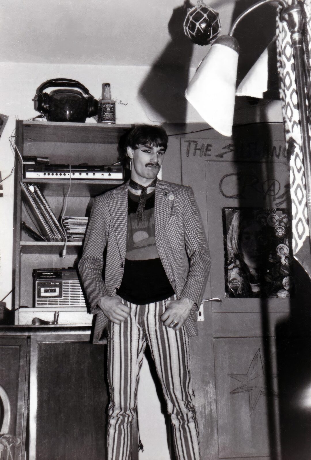 A man with a mustache stands indoors, wearing a blazer, striped pants, and a graphic t-shirt. Behind him are shelves with records, a radio, and various objects. The image is in black and white.